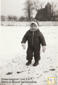 Czarno-biała fotografia z lat 80. XX wieku. Małe dziecko ubrane w gruby zimowy kombinezon, czapkę, szalik i rękawiczki stoi na ośnieżonym polu. W tle widoczny jest kościół filialny pw. św. Jerzego w Przyczynie Górnej oraz bezlistne drzewa. Zimowy krajobraz wsi.