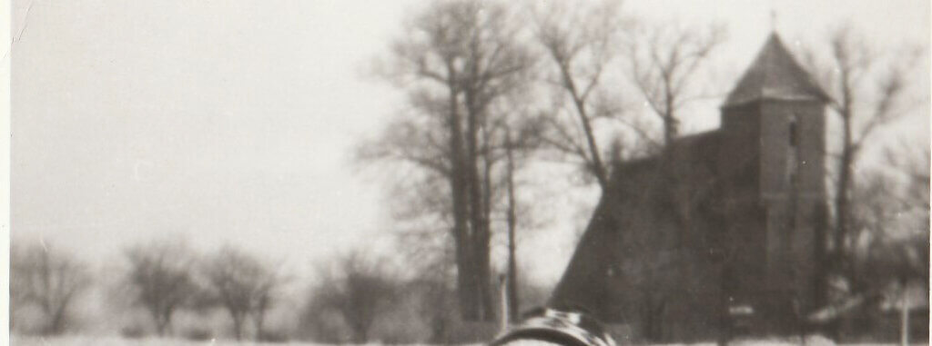 Czarno-biała fotografia z lat 80. XX wieku. Zimowy krajobraz wsi: ośnieżone pole, bezlistne drzewa oraz kościół filialny pw. św. Jerzego w Przyczynie Górnej widoczny w tle.