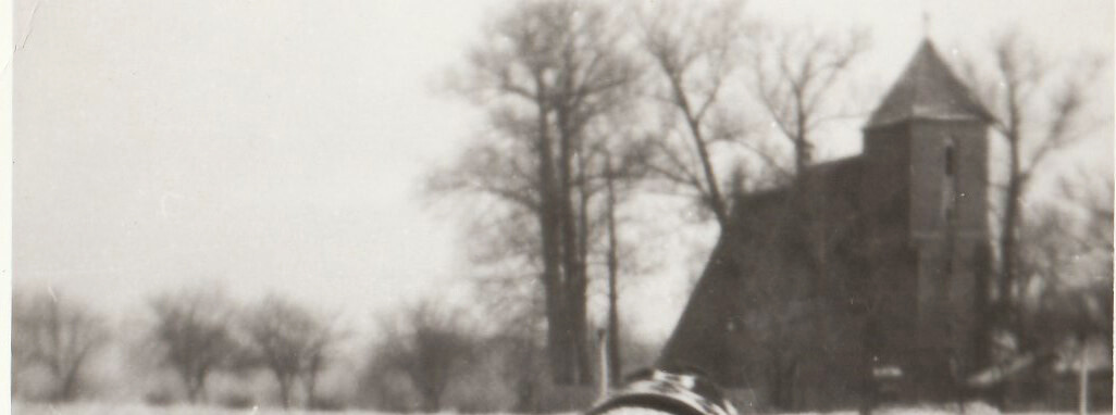 Czarno-biała fotografia z lat 80. XX wieku. Zimowy krajobraz wsi: ośnieżone pole, bezlistne drzewa oraz kościół filialny pw. św. Jerzego w Przyczynie Górnej widoczny w tle.