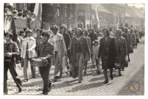 Czarno-biała fotografia przedstawiająca pochód uliczny z lat 70. XX wieku. Na brukowanej ulicy maszeruje grupa kobiet, dzieci oraz harcerzy, w tym chłopiec grający na bębnie na czele kolumny. W tle widać tłum obserwatorów stojących na podwyższeniu oraz zabudowę miejską z kamienicami. Wzdłuż ulicy rozmieszczone są flagi i dekoracje. Zdjęcie pochodzi ze zbiorów prywatnych, udostępnione przez Stowarzyszenie Czas A.R.T.
