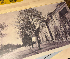 Reprodukcja archiwalnej, czarno-białej fotografii przedstawiającej budynek Starostwa Powiatowego we Wschowie w latach 30. XX wieku. Budynek stoi przy szerokiej, wysadzanej drzewami ulicy. Widać elewację z licznymi oknami oraz charakterystyczną architekturę okresu międzywojennego