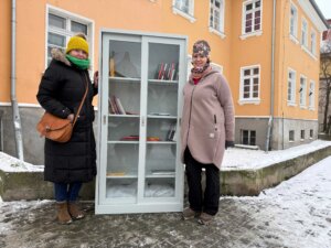Dwie kobiety stoją na zewnątrz, zimą, po obu stronach przeszklonej szafy bookcrossingowej wypełnionej książkami. Jedna ma na sobie czarny płaszcz, żółtą czapkę i zielony szalik, druga jasny płaszcz i wzorzystą czapkę. Szafa stoi przy jasnopomarańczowym budynku z białymi oknami, na chodniku pokrytym śniegiem. Zdjęcie dokumentuje uruchomienie punktu wymiany książek.