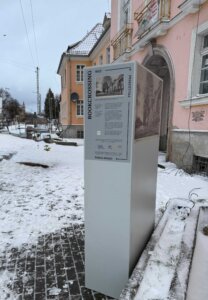 Przeszklona szafa do wymiany książek stoi na zewnątrz, na zaśnieżonym chodniku, przed historycznym budynkiem z jasnoróżową elewacją i kamiennym portalem wejściowym. Na boku szafy widoczna jest plansza informacyjna z napisem »Bookcrossing«, archiwalnym zdjęciem budynku oraz instrukcjami: »Weź«, »Przeczytaj«, »Podaj dalej«. Zdjęcie pokazuje punkt wymiany książek w przestrzeni publicznej.