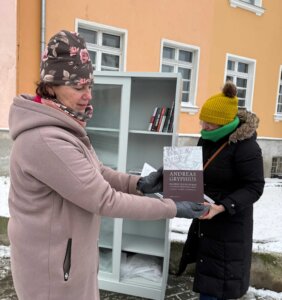 Dwie kobiety stoją na zewnątrz przy przeszklonej szafie wymiany książek. Jedna z nich trzyma w rękach książkę i przekazuje ją drugiej osobie. Obie są ubrane w zimowe płaszcze, czapki i rękawiczki. W tle widoczna jest jasna elewacja budynku oraz szafa bookcrossingowa z półkami na książki. Zdjęcie pokazuje moment dzielenia się książką.