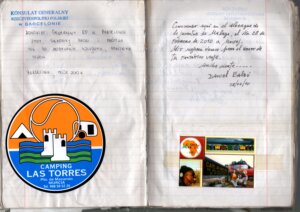 Zdjęcie przedstawia dwie otwarte strony podróżniczego notesu. Kartki są jasne, lekko zniszczone i zapisane ręcznie. Na górze lewej strony widnieje niebieski nadruk „Konsulat Generalny Rzeczypospolitej Polskiej w Barcelonie” oraz odręczne notatki w języku polskim, z datą z 2010 roku. Na górze prawej strony znajduje się ręcznie napisany tekst w języku hiszpańskim – osobista wiadomość z życzeniami powodzenia w dalszej podróży, podpisana imieniem i nazwiskiem autora oraz datą. W dolnej części lewej strony umieszczona jest duża, okrągła, kolorowa naklejka z logo „Camping Las Torres” z Murcji, przedstawiająca stylizowany zamek, góry i fale. W dolnej części prawej strony znajdują się cztery małe fotografie: kolorowe domki, krajobraz, portret uśmiechniętego mężczyzny oraz zdjęcie podróżników przy tablicy „Cape of Good Hope”. Całość ma charakter osobistego archiwum podróży, łączącego notatki, pieczątki i wizualne pamiątki z drogi.