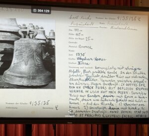 Czarno-biała archiwalna fotografia XVIII-wiecznego dzwonu z 1730 roku odlanego przez Stephana Wernera w Lesznie, prezentowana na ekranie podczas wykładu w muzeum. Obok zdjęcia widoczna jest karta inwentarzowa z odręcznymi notatkami w języku niemieckim, zawierająca dane techniczne dzwonu, jego wymiary, materiał (brąz), miejsce odlania oraz opis dekoracji i inskrypcji. Ekran stoi na tle czerwonych kotar w sali muzealnej.