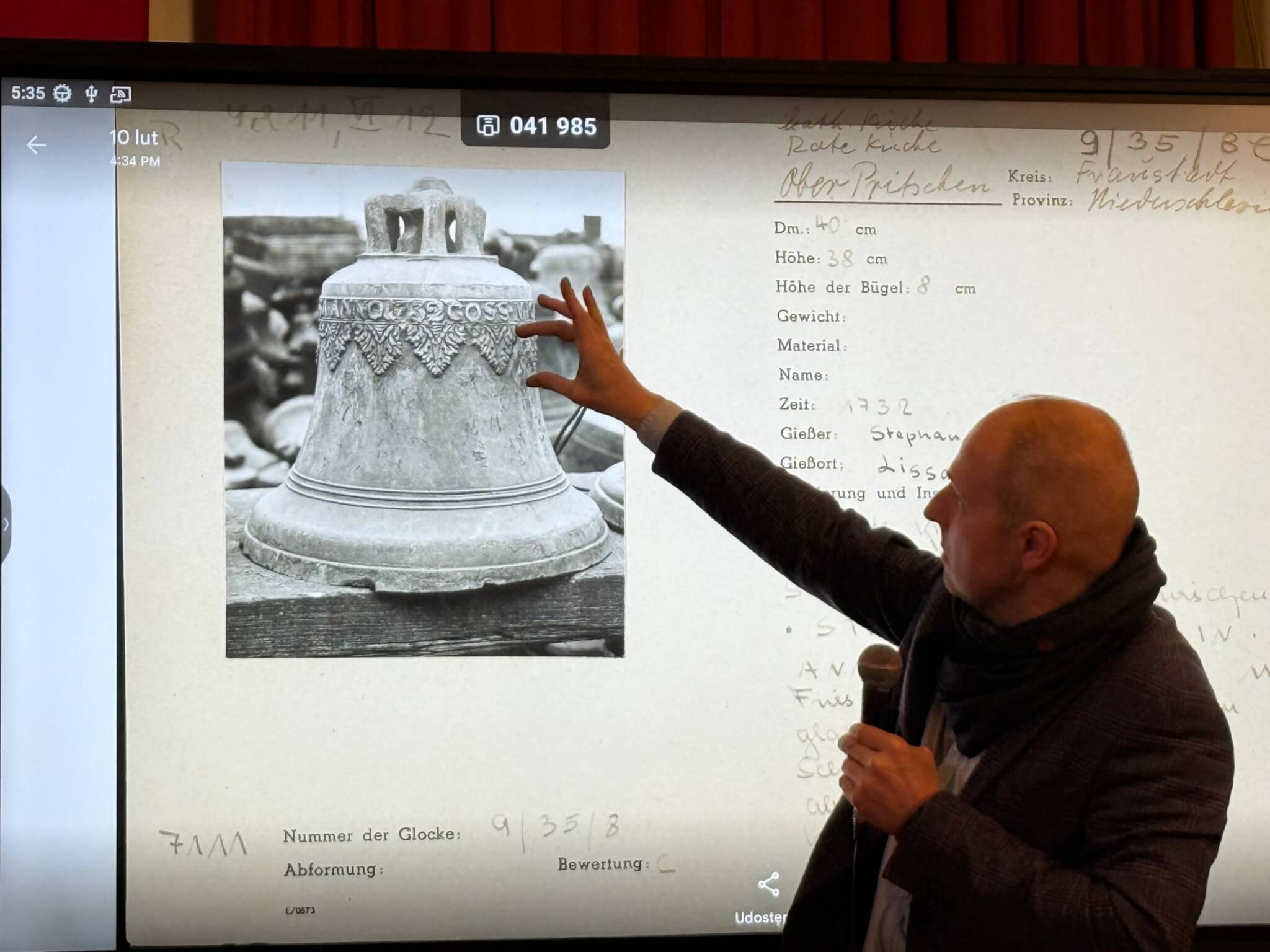 Prelegent podczas wykładu w sali muzealnej wskazuje na ekran multimedialny, na którym wyświetlona jest archiwalna, czarno-biała fotografia XVIII-wiecznego dzwonu autorstwa Stephana Wernera. Obok zdjęcia widoczna jest karta dokumentacyjna z opisem technicznym obiektu. Mężczyzna trzyma mikrofon i omawia detale dekoracji dzwonu, a w tle widać czerwone kotary i fragment ekspozycji muzealnej.