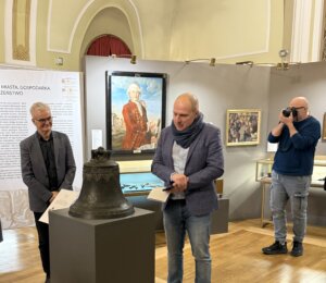 W sali muzealnej podczas prezentacji nowego nabytku prelegent stoi obok XVIII-wiecznego dzwonu ustawionego na postumencie i omawia jego historię. Obok niego stoi drugi uczestnik wydarzenia z dokumentami w ręku, a w tle widoczne są obrazy i gabloty ekspozycyjne. Po prawej stronie fotograf dokumentuje spotkanie. Sala ma jasne ściany, drewnianą podłogę i muzealną aranżację wystawienniczą.
