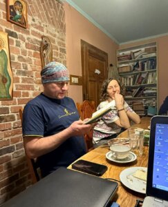 Zdjęcie przedstawia spotkanie przy drewnianym stole w domowym wnętrzu. Po lewej stronie siedzi mężczyzna w chuście na głowie i ciemnej koszulce, który czyta lub przegląda zeszyt z notatkami. Obok niego siedzi kobieta, oparta o stół, patrząca w jego stronę z uśmiechem i dłonią przy twarzy. Na stole znajdują się filiżanka, szklanki, talerz z kawałkiem ciasta, telefon oraz zamknięty laptop. Po prawej stronie kadru widać fragment otwartego laptopa. W tle jest ceglana ściana z ikonami, drewniane drzwi oraz regał z książkami. Atmosfera zdjęcia jest spokojna i kameralna, sugerująca rozmowę lub wspólne czytanie wspomnień.