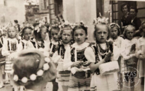 Czarno-biała archiwalna fotografia przedstawiająca procesję Bożego Ciała we Wschowie w 1954 roku. Na pierwszym planie idzie grupa kilkuletnich dziewczynek ubranych w krakowskie stroje ludowe z wiankami z kwiatów na głowach i koralami na szyjach. Dzieci niosą małe koszyczki i świece, idąc skupione w szeregu środkiem ulicy. W tle widoczne są kolejne uczestniczki procesji oraz zabudowa miasta. Druga dziewczynka od prawej to Elżbieta Lewandowska, za nią stoi Janina Nowacka. Zdjęcie dokumentuje powojenne życie religijne i społeczne mieszkańców Wschowy.