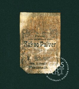 Zniszczona, pożółkła etykieta opakowania kakao z przełomu XIX i XX wieku odnaleziona w kamienicy. Na środku widoczny ozdobny napis w języku niemieckim „Kakao-Pulver” otoczony dekoracyjną ramką. Poniżej czytelny fragment nazwy właściciela lub sklepu: "E. Fengler, Fraustadt”. Papier jest silnie zabrudzony, popękany i nadpalony, z ubytkami w dolnej części. Na etykiecie widoczny znak wodny „CZAS A.R.T.”.