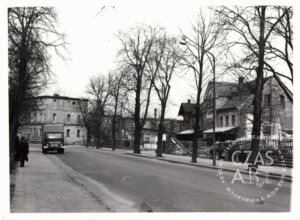 Archiwalne czarno-białe zdjęcie ulicy Kazimierza Wielkiego we Wschowie z lat 70. XX wieku. Po prawej dawna rozlewnia GS i zabudowania browaru, po lewej ciężarówka i przechodnie na chodniku, wzdłuż ulicy rząd wysokich drzew.