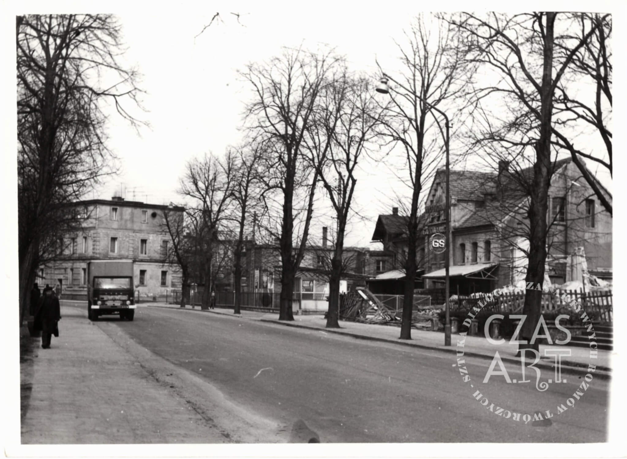 Archiwalne czarno-białe zdjęcie ulicy Kazimierza Wielkiego we Wschowie z lat 70. XX wieku. Po prawej dawna rozlewnia GS i zabudowania browaru, po lewej ciężarówka i przechodnie na chodniku, wzdłuż ulicy rząd wysokich drzew.