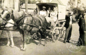 Czarno-biała archiwalna fotografia z końca lat 50. XX wieku we Wschowie. Na pierwszym planie stoi zaprzęg konny z drewnianym wozem przykrytym płócienną budą. Na koźle siedzi woźnica – Onufry Kubicki – a obok niego Matylda Szymczakowska z córką. W wozie widoczne są osoby jadące na plener malarski. Po prawej stronie stoją dwaj mężczyźni przyglądający się scenie. W tle widoczna zabudowa i drzewa. Zdjęcie dokumentuje wyjazd członków Klubu Twórców Amatorów z Domu Kultury we Wschowie na zajęcia plenerowe.