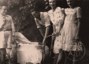 Czarno-biała, archiwalna fotografia z lat powojennych. Na podwórku lub w ogrodzie stoją cztery osoby: mężczyzna pochylony nad jasnym dziecięcym wózkiem oraz troje młodych ludzi — dwie dziewczyny i chłopiec. Jedna z dziewczyn trzyma dłonie na rączce wózka, druga stoi obok, uśmiechnięta do obiektywu. Chłopiec po lewej stronie opiera się swobodnie, również patrząc w stronę fotografa. W tle widoczna gęsta roślinność i liście drzew. Zdjęcie ma charakter rodzinny i dokumentuje codzienną scenę z życia powojennej Wschowy.