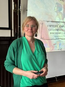 Kobieta prowadząca wykład stoi przed ekranem projekcyjnym, trzyma pilot do prezentacji.