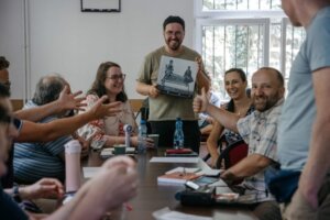 Grupa osób siedzi przy stole podczas spotkania lub warsztatów. W centrum stoi uśmiechnięty mężczyzna, który prezentuje książkę lub publikację. Pozostali uczestnicy reagują pozytywnie – część bije brawo, ktoś pokazuje kciuk w górę. Na stole widać notatki, butelki z wodą i materiały. Atmosfera jest swobodna i życzliwa.