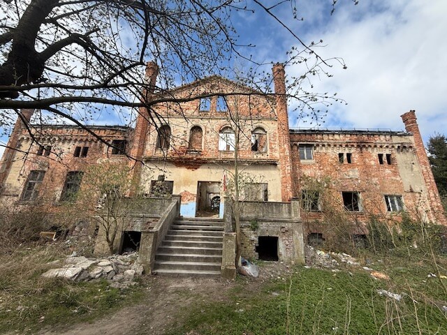 Front opuszczonego, zrujnowanego budynku z czerwonej cegły z centralnymi schodami prowadzącymi do wejścia; wybite okna, uszkodzona elewacja i roślinność porastająca teren wokół.