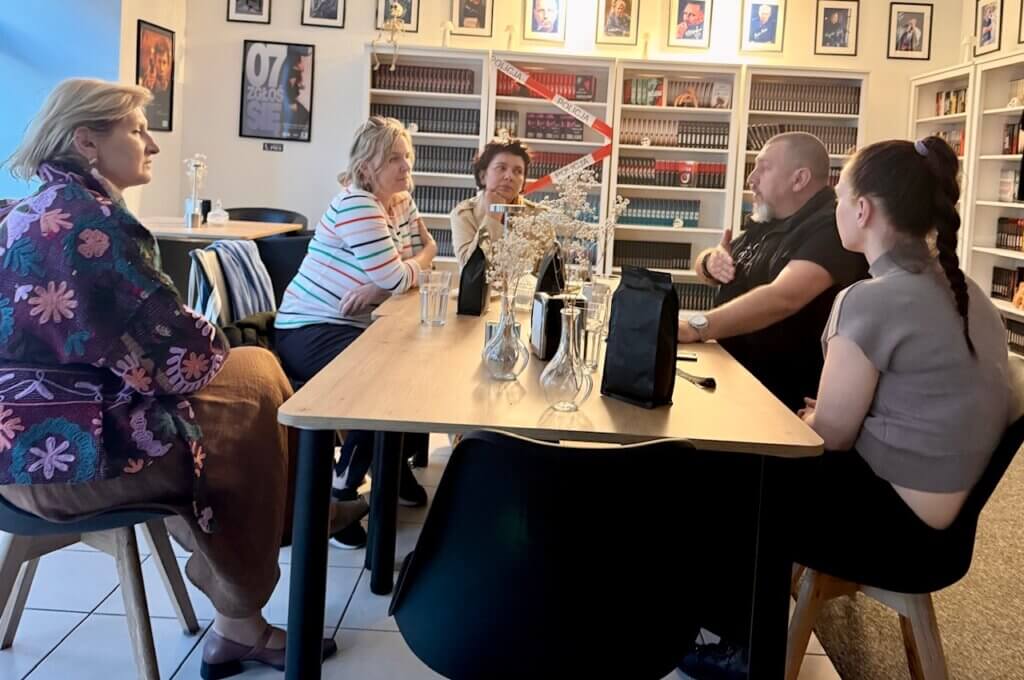 Grupa pięciu osób siedzi przy stole w kawiarni otoczonej regałami z książkami. Na stole stoją szklanki z wodą i opakowania kawy, jedna osoba gestykuluje podczas rozmowy.