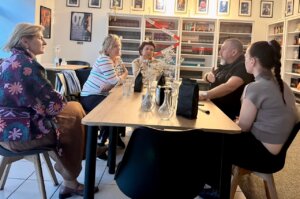 Grupa pięciu osób siedzi przy stole w kawiarni otoczonej regałami z książkami. Na stole stoją szklanki z wodą i opakowania kawy, jedna osoba gestykuluje podczas rozmowy.