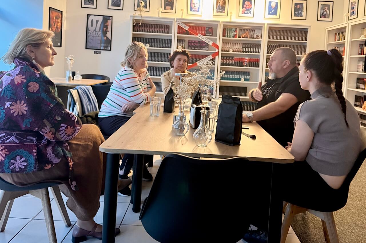 Grupa pięciu osób siedzi przy stole w kawiarni otoczonej regałami z książkami. Na stole stoją szklanki z wodą i opakowania kawy, jedna osoba gestykuluje podczas rozmowy.