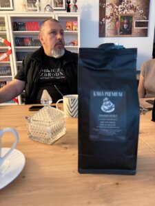 Mężczyzna siedzi przy stole w kawiarni, w tle regały z książkami i obrazy na ścianie. Na stole widoczne są filiżanki i opakowanie kawy na pierwszym planie.