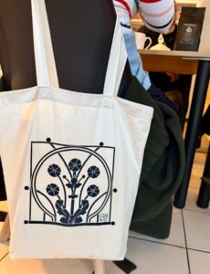 Materiałowa torba w kolorze jasnym zawieszona na krześle. Na torbie widoczny czarny, roślinny wzór graficzny oraz napis „Czas A.R.T.”.