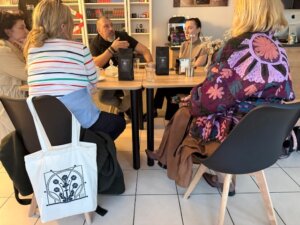 Kilka osób siedzi przy stolikach ustawionych razem i rozmawia w przestrzeni z książkami. Na oparciu krzesła wisi materiałowa torba z graficznym wzorem.