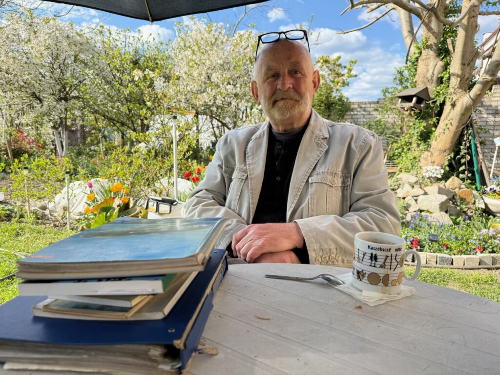 Mężczyzna siedzi przy stole w ogrodzie, przed nim leży stos starych albumów i dokumentów. Ma siwe włosy i brodę, ubrany jest w jasną kurtkę i czarną koszulę, na głowie opiera okulary. Na stole stoi kubek z napisem „Kaszëbsczé nutë” oraz łyżeczka. W tle widoczny ogród z kwitnącymi drzewami, kwiatami i kamiennym murkiem.