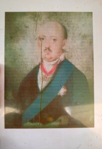 Kolorowy portret mężczyzny w eleganckim stroju z odznaczeniami i niebieską szarfą, z widocznym uszkodzeniem fotografii w postaci pionowego pęknięcia.