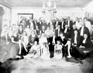 Zdjęcie grupowe (prawdopodobnie wesele lub uroczystość) Archiwalne czarno-białe zdjęcie dużej grupy osób w eleganckich strojach, ustawionych we wnętrzu z żyrandolem. W centrum siedzi panna młoda z bukietem.