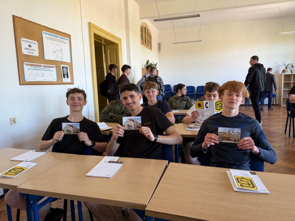 Uczniowie siedzący w szkolnych ławach. Trzej uczniowie w pierwszej ławce pokazują otrzymanej kartki pocztowe przedstawiajacą kamienicę w mieście Wschowa