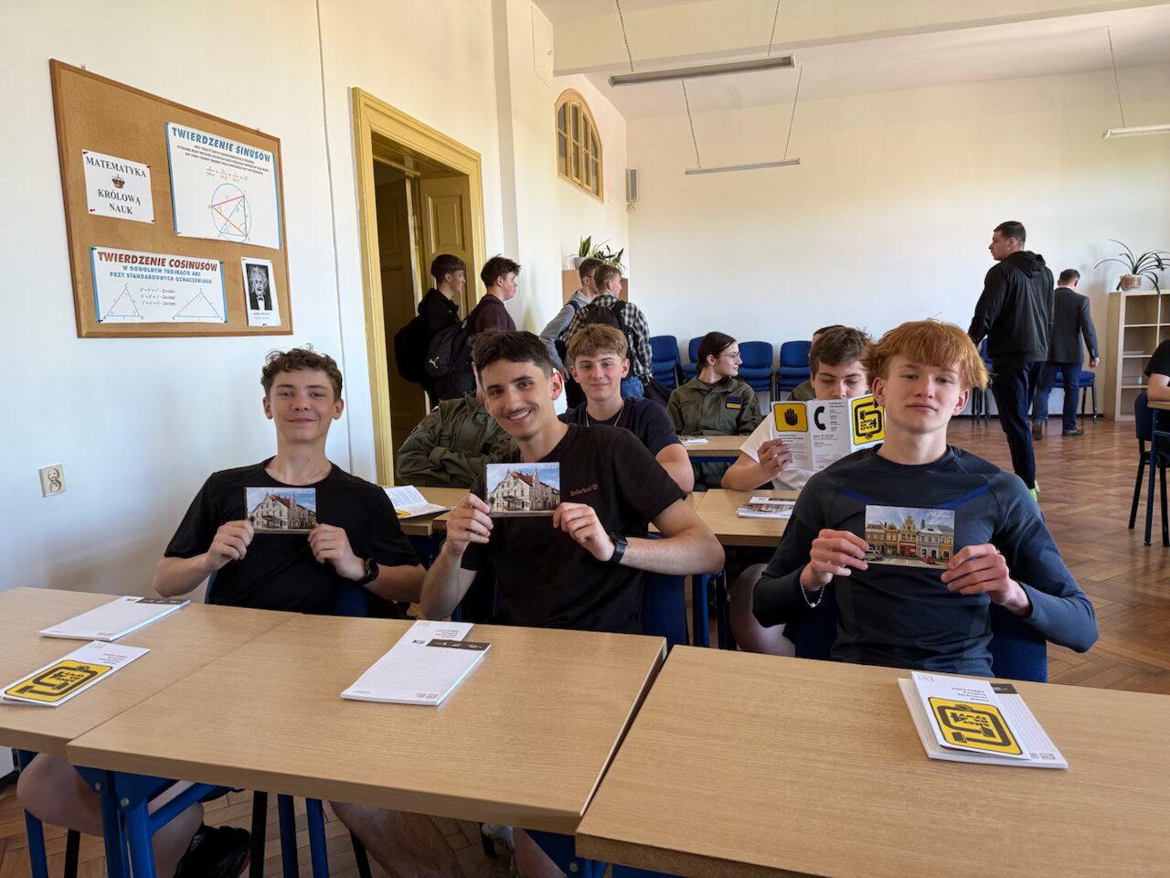 Uczniowie siedzący w szkolnych ławach. Trzej uczniowie w pierwszej ławce pokazują otrzymanej kartki pocztowe przedstawiajacą kamienicę w mieście Wschowa