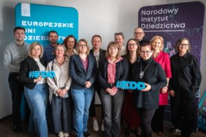 Grupa kilkunastu osób stoi w pomieszczeniu konferencyjnym między dwoma roll-upami z napisami „Europejskie Dni Dziedzictwa” i „Narodowy Instytut Dziedzictwa”; część osób trzyma niebieskie napisy „#EDD”.