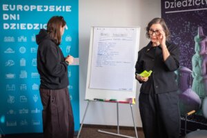 Dwie osoby stoją przy flipcharcie z zapisanymi punktami; jedna z nich trzyma karteczki i mówi, druga słucha; w tle baner „Europejskie Dni Dziedzictwa”.