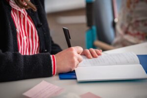 Zbliżenie dłoni zapisującej notatki w otwartym zeszycie przy użyciu czarnego długopisu; na stole widoczne są karteczki i materiały.