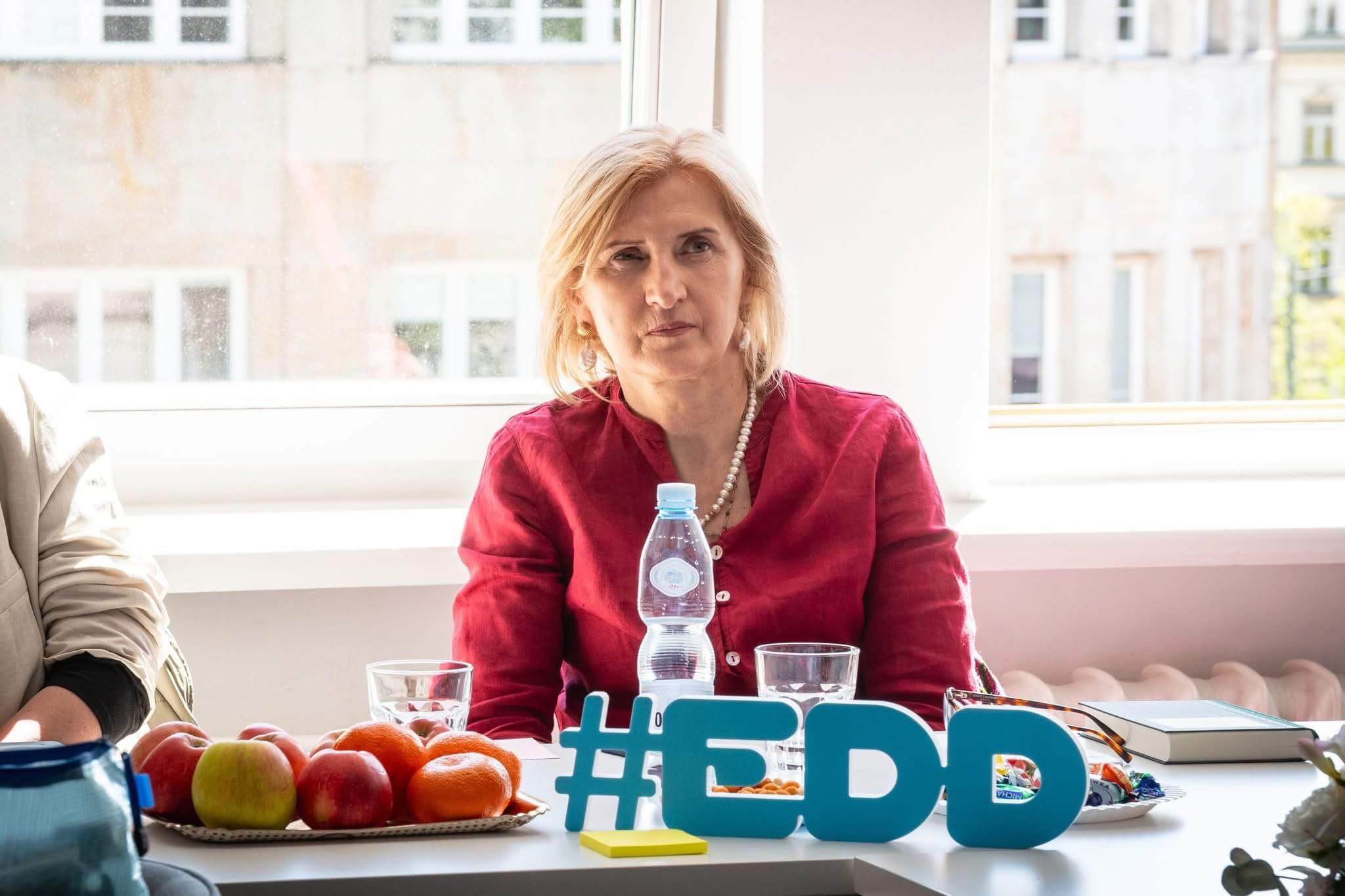 Kobieta siedzi przy stole konferencyjnym, przed nią stoi butelka wody, owoce i niebieski napis „#EDD”, w tle jasne okno.