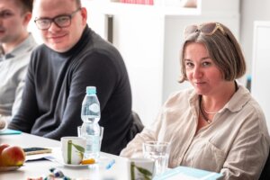 Dwie osoby siedzą przy stole konferencyjnym z kubkami, wodą i notatkami; jedna patrzy w stronę aparatu, druga lekko się uśmiecha.