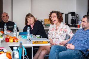 Kilka osób siedzi przy stole konferencyjnym, rozmawia i reaguje na wypowiedź; na stole widoczne są książki, owoce, kubki i materiały szkoleniowe.