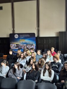 Grupa uczniów słuchająca wykładu, w tle widoczny baner szkoły „ZAN – trampolina do przyszłości”.