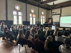 Widok sali wykładowej – uczestnicy siedzą na krzesłach i oglądają prezentację wyświetlaną na ekranie.