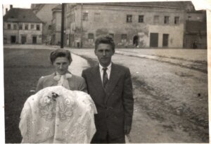 Czarno-biała fotografia młodej pary stojącej na ulicy miasta. Kobieta trzyma ozdobnie haftowaną tkaninę lub szatę, obok niej stoi mężczyzna w garniturze i krawacie. W tle widoczne są stare kamienice oraz brukowana droga.