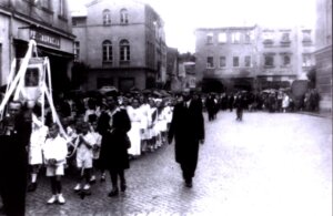 Czarno-białe zdjęcie przedstawiające procesję lub uroczysty pochód na brukowanej ulicy starego miasta. Po lewej stronie stoją dzieci ubrane na biało, a środkiem ulicy idą mieszkańcy w odświętnych strojach. W tle widoczne są zabytkowe kamienice i zniszczona zabudowa.