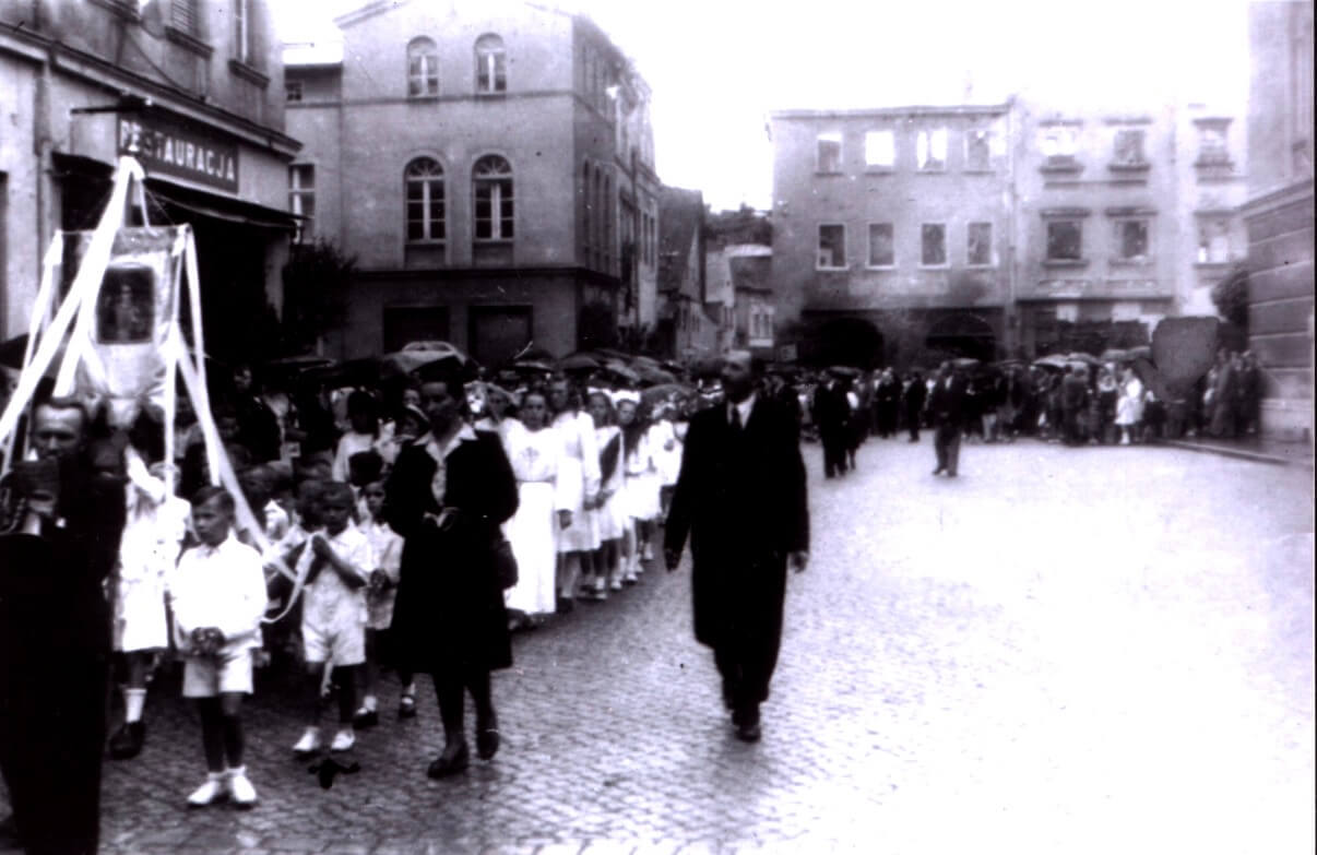 Czarno-białe zdjęcie przedstawiające procesję lub uroczysty pochód na brukowanej ulicy starego miasta. Po lewej stronie stoją dzieci ubrane na biało, a środkiem ulicy idą mieszkańcy w odświętnych strojach. W tle widoczne są zabytkowe kamienice i zniszczona zabudowa.