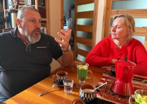 Kolorowe zdjęcie przedstawiające dwie osoby siedzące przy stole w pokoju. W tle regał z książkami. Na stole filiżanki z kawą i imbryk do kawy. Osoby widoczny na zdjęciu po lewej stronie mężczyzna po prawej stronie kobieta rozmawiają ze sobą.