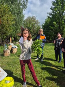 Kolorowa fotografia na której widać panią stojącą tłumaczącą coś zebranym osobą trzymająca w ręku kwiaty z pola