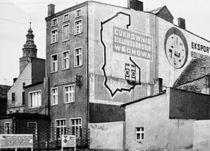 Czarno-biała fotografia przedstawiająca fragment zabudowy Wschowy z lat 70. XX wieku. Na bocznej ścianie kamienicy widoczne jest wielkoformatowe malowidło reklamowe z napisem „Cukrownie Zielonogórskie Wschowa”, konturem województwa oraz symbolem buraka cukrowego. Po prawej stronie znajduje się fragment reklamy „Eksport Rolimpex”. W tle widoczna jest wieża kościoła. Fotografia dokumentuje charakterystyczne elementy przestrzeni miejskiej z okresu PRL oraz miejsce, w którym przez wiele lat funkcjonował wschowski skansen. Zdjęcie zostało opublikowane w ramach konkursu „Ocalmy od zapomnienia 2026”, poświęconego dokumentowaniu historii dawnej ziemi wschowskiej.