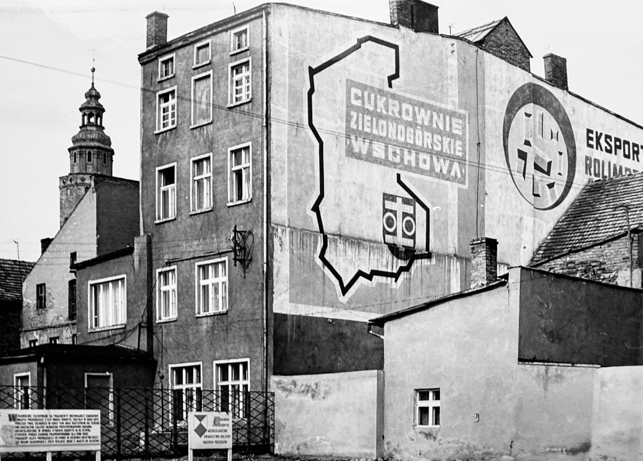Czarno-biała fotografia przedstawiająca fragment zabudowy Wschowy z lat 70. XX wieku. Na bocznej ścianie kamienicy widoczne jest wielkoformatowe malowidło reklamowe z napisem „Cukrownie Zielonogórskie Wschowa”, konturem województwa oraz symbolem buraka cukrowego. Po prawej stronie znajduje się fragment reklamy „Eksport Rolimpex”. W tle widoczna jest wieża kościoła. Fotografia dokumentuje charakterystyczne elementy przestrzeni miejskiej z okresu PRL oraz miejsce, w którym przez wiele lat funkcjonował wschowski skansen. Zdjęcie zostało opublikowane w ramach konkursu „Ocalmy od zapomnienia 2026”, poświęconego dokumentowaniu historii dawnej ziemi wschowskiej.